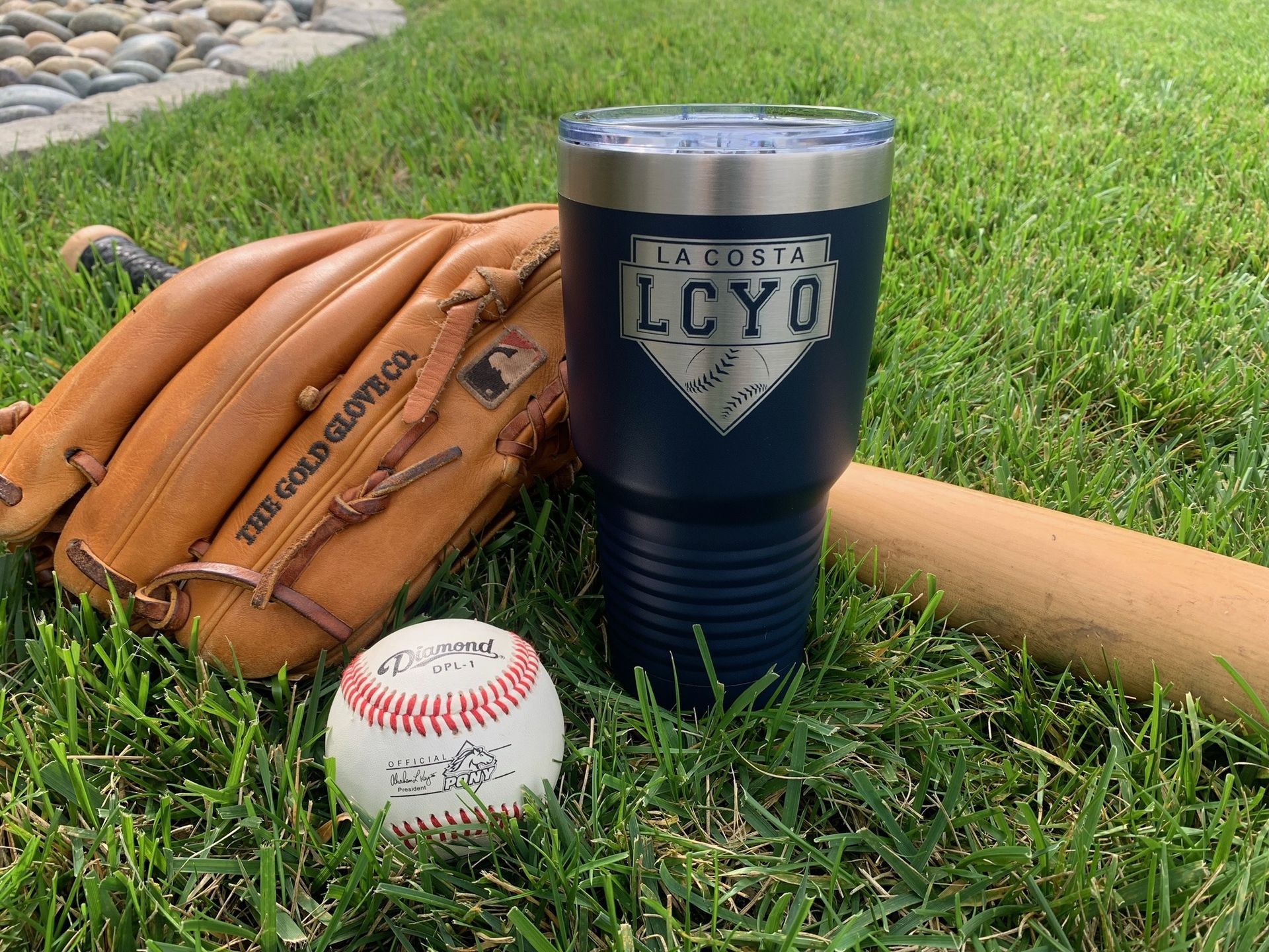 Baseball glove, ball, bat, and navy tumbler on grass near a stone border
