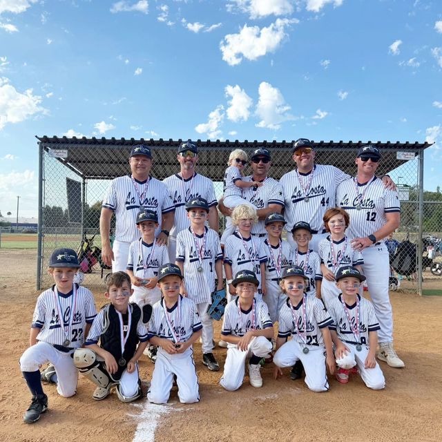 Youth baseball team and coaches posing on a dirt field in front of the dugout, holding a championship trophy.