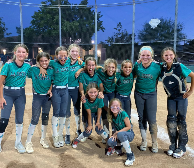 Youth softball team in teal jerseys posing together on a dirt field at dusk, smiling after a game.