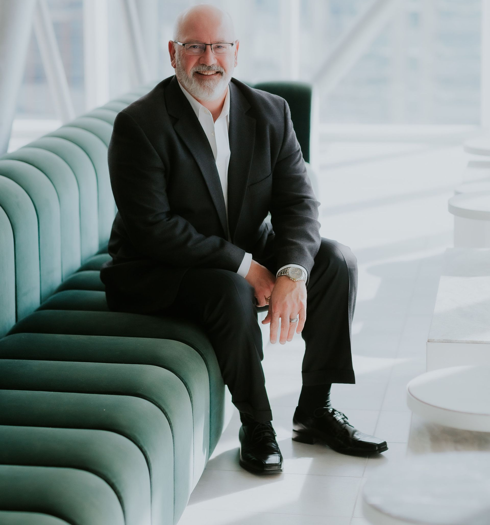 Man in a suit smiles, seated on a green, curved bench in a modern space.