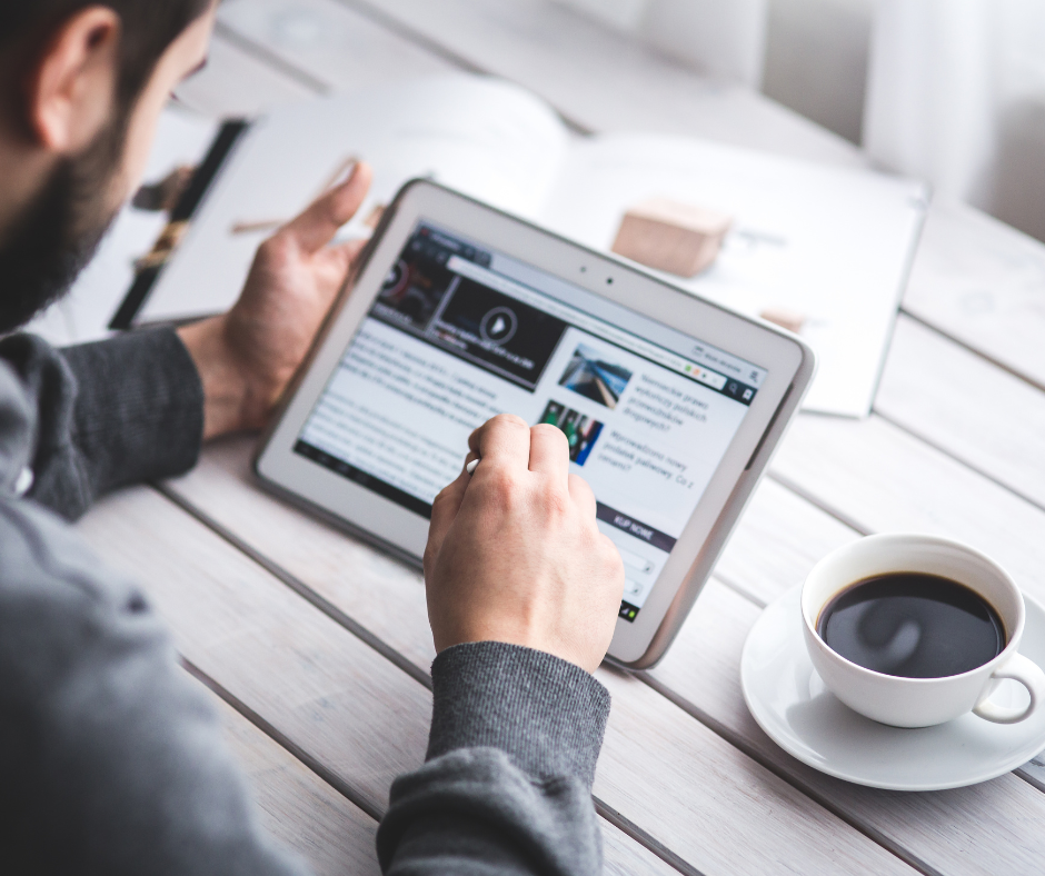 Person using a tablet at a white table, holding a pen. A cup of coffee sits beside the tablet.