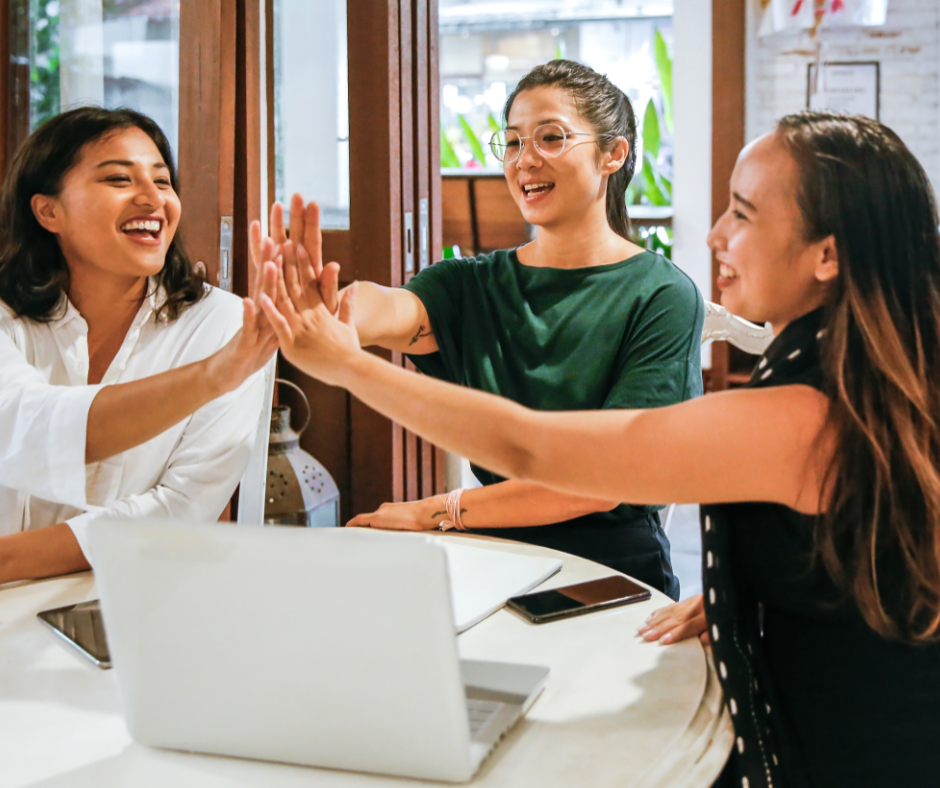 Three women smiling, high-fiving over a laptop at a table, sunny interior.