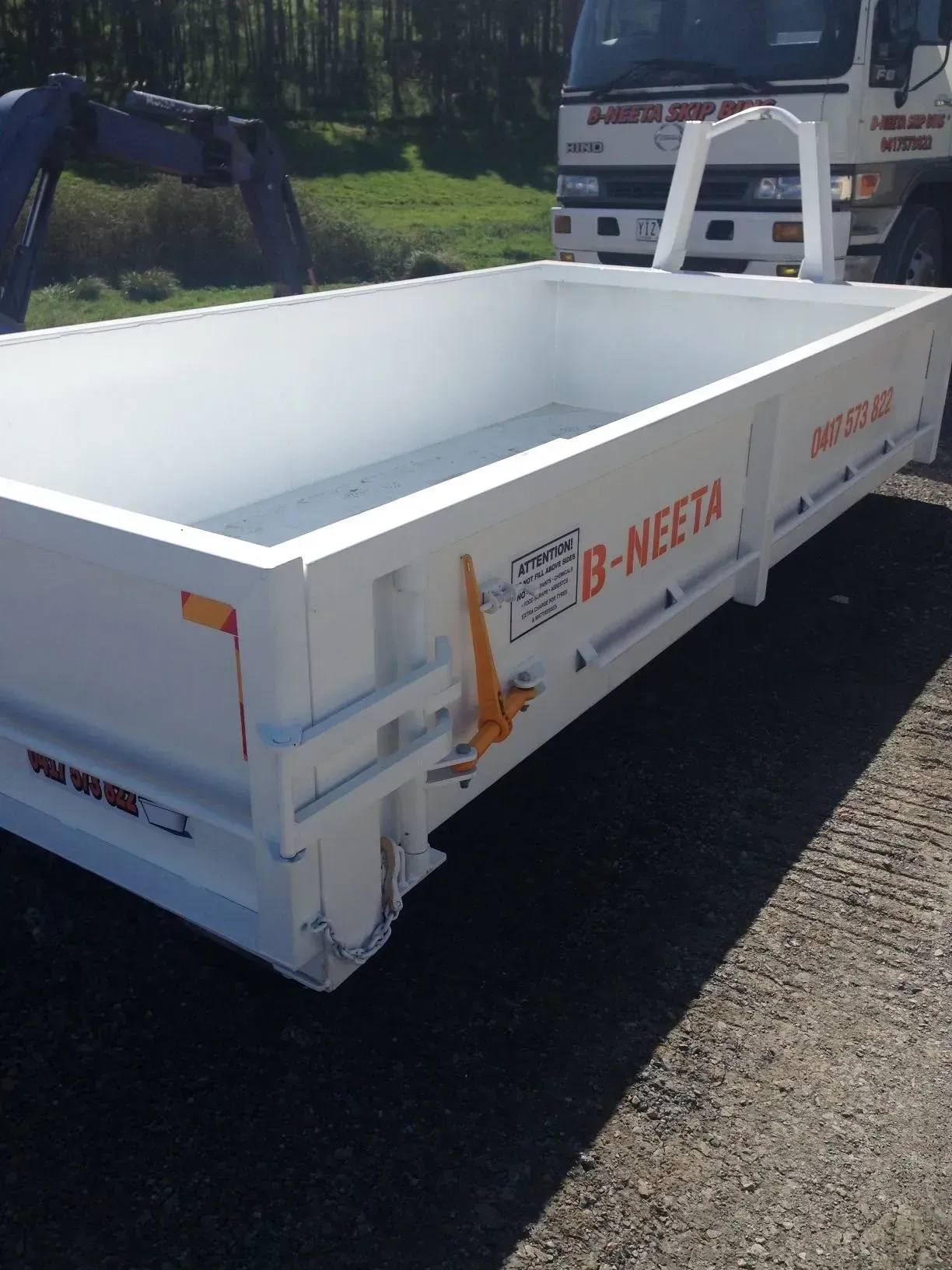White open-top dumpster container on a truck bed, parked on a road with a backhoe in the background