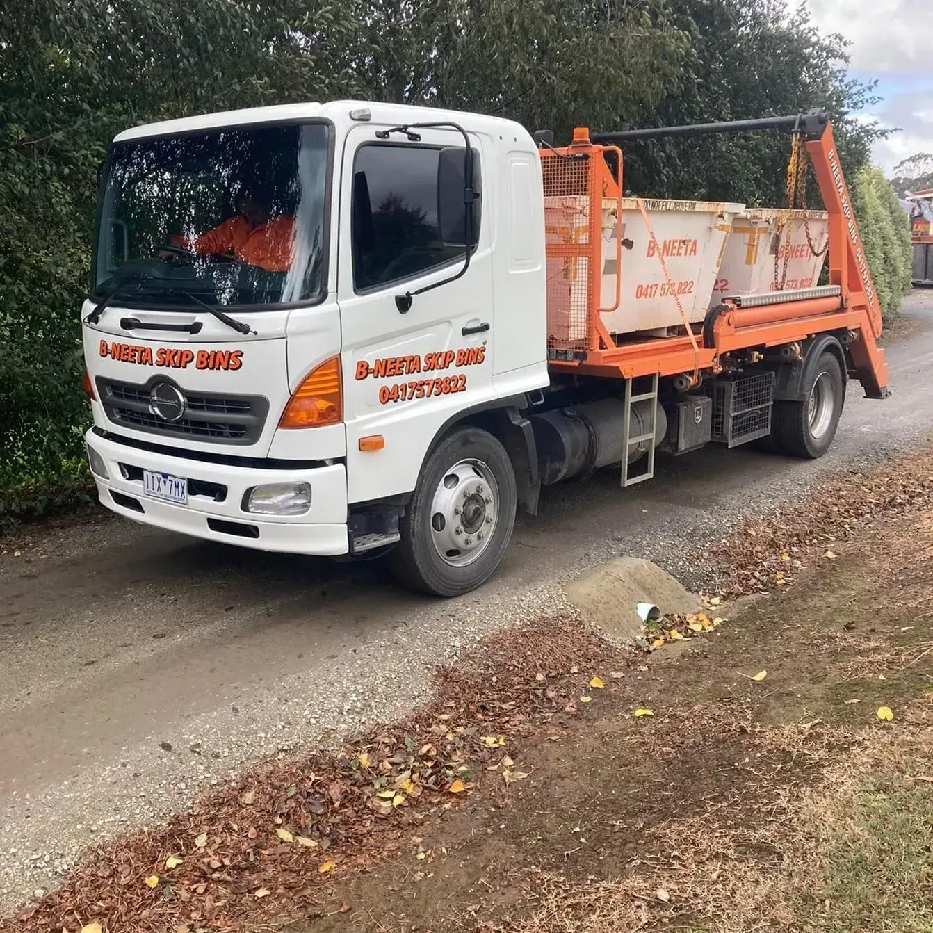 White and orange utility truck on a narrow rural road with trees and fallen leaves nearby