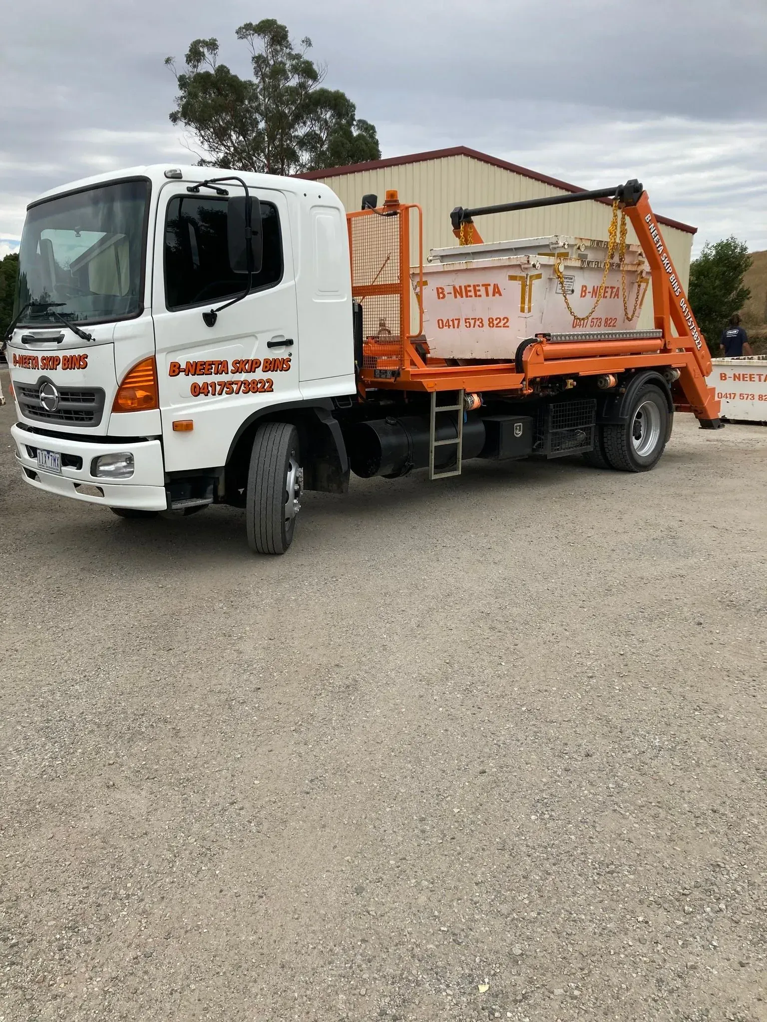 Orange-and-white garbage truck parked on gravel in front of a building and trees