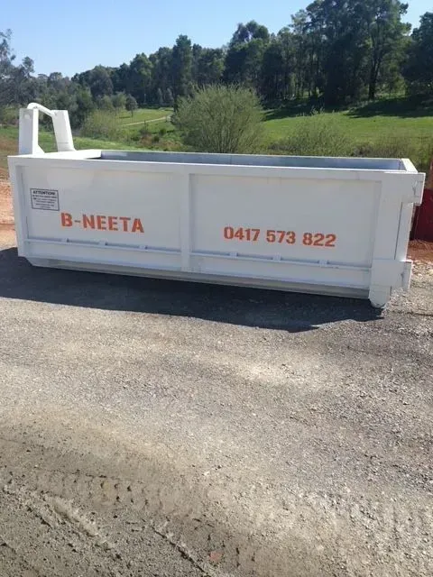 White B-NEETA skip bin with orange lettering on a roadside, with green trees in the background