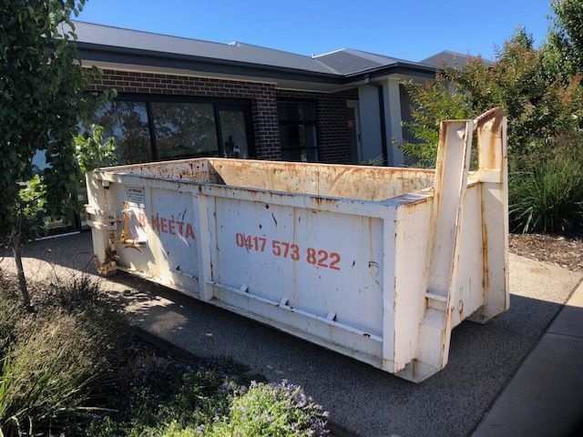 Large white skip bin with orange writing in a driveway beside a brick house and garden