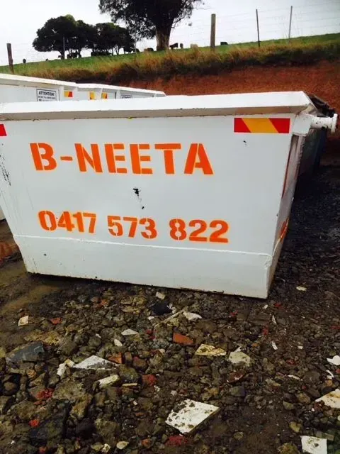 White B-NEETA dumpster with orange lettering and phone number, on rocky ground outdoors