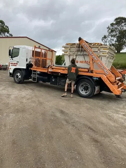 Orange flatbed truck with ladder equipment parked on a dirt lot, with a worker standing beside it
