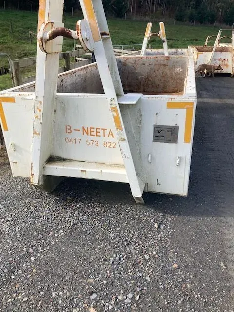 White skip bin on gravel with orange markings and a strap across the front, set outdoors near trees.