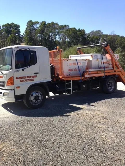 White truck with an orange dumpster bin parked on gravel under a blue sky.