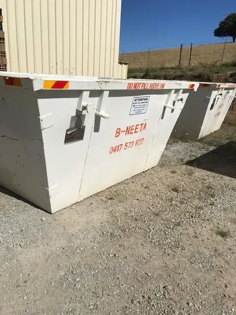 White utility dumpster/bin with orange warning text beside a beige building on gravel, under a clear sky