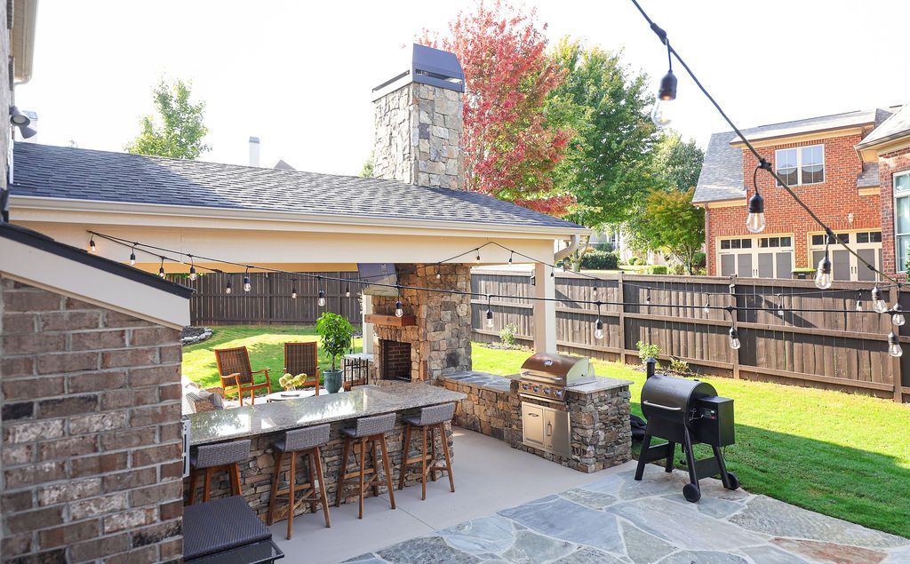 Covered backyard patio with bar stools, grill, string lights, and a fenced garden area.
