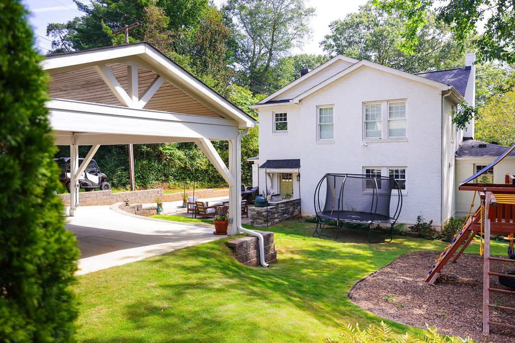 Backyard with a white house, wooden carport, green lawn, and a playset on the right
