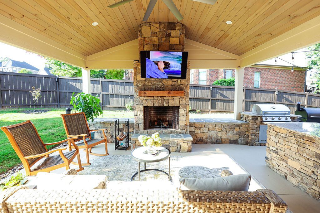Covered outdoor patio with stone fireplace, TV, seating chairs, and stone bar under a wood ceiling