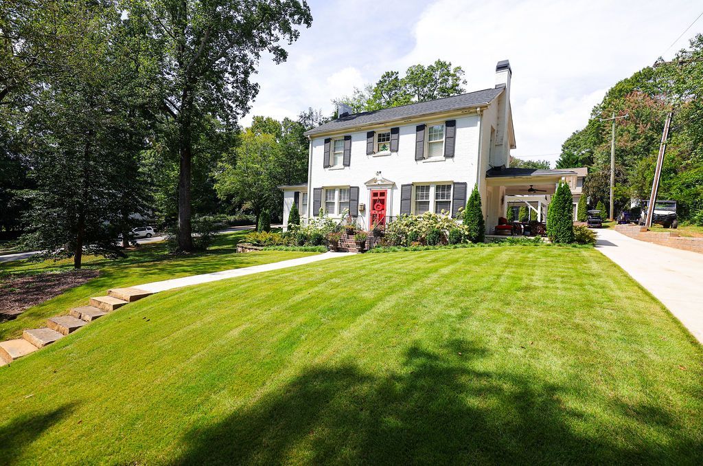 White house with red door on a green lawn beside a tree-lined road on a sunny day