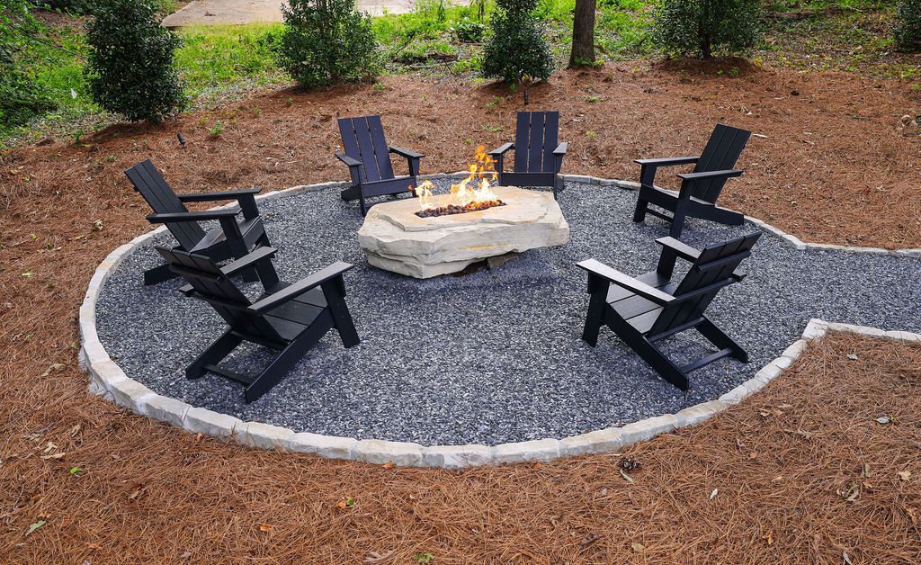 Outdoor fire pit seating area with six black chairs around a stone fire pit on a gravel patio.