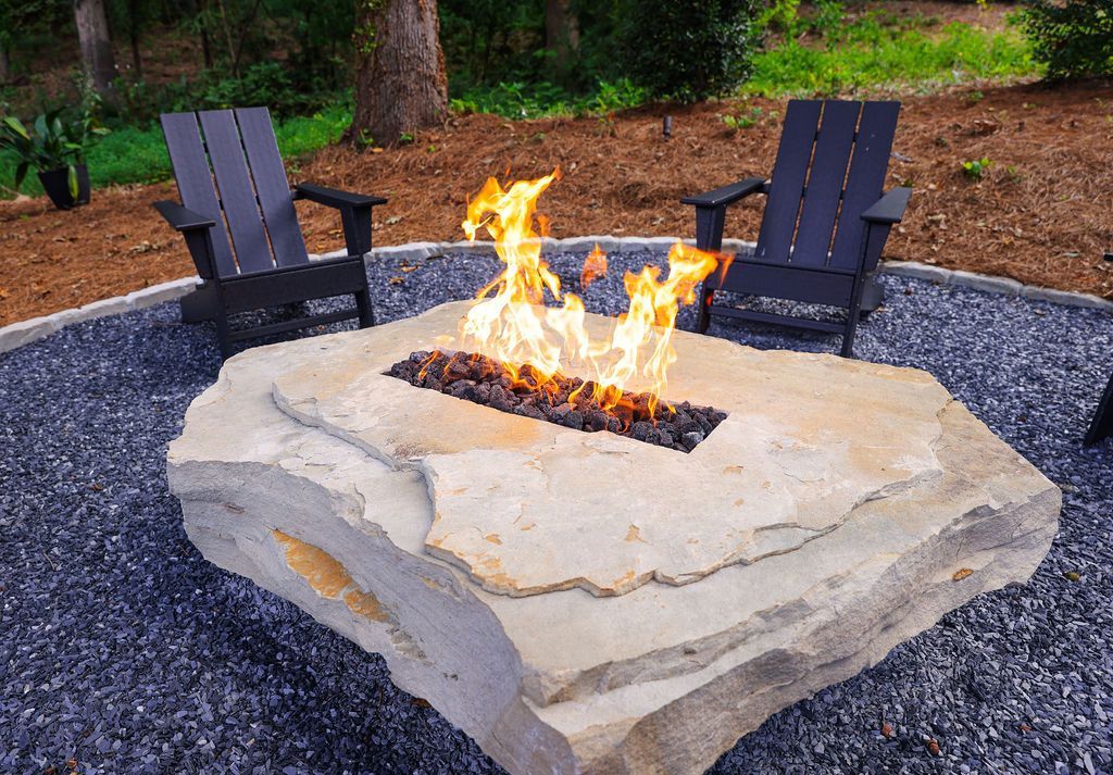 Stone fire pit with flames, surrounded by two dark lounge chairs on a gravel patio outdoors
