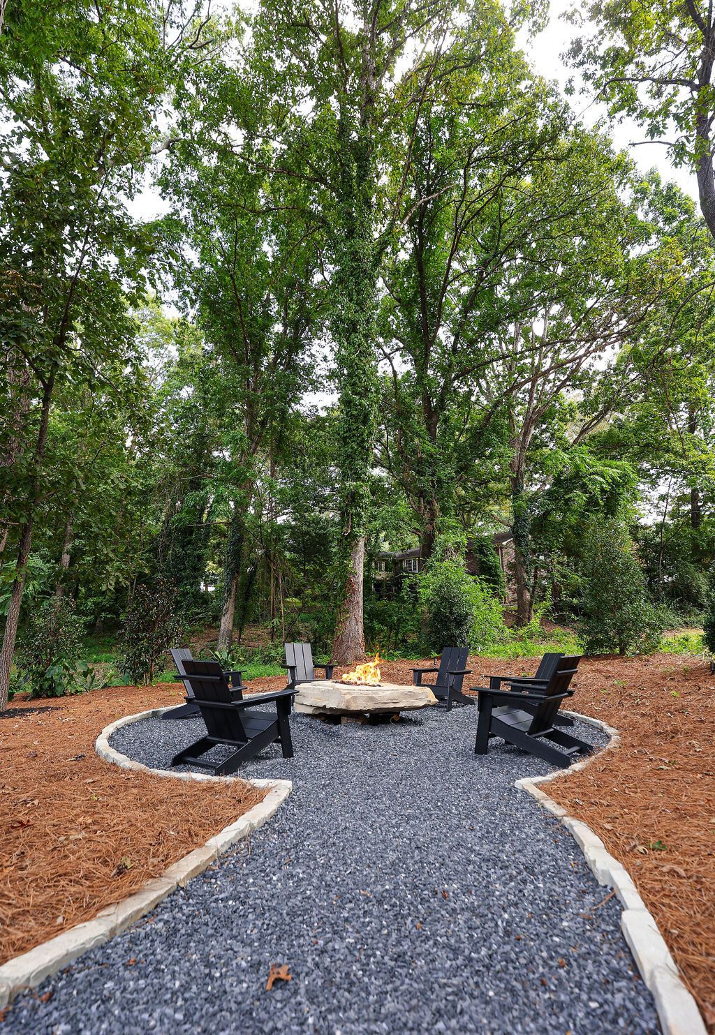 Gravel path leading to a fire pit with black chairs in a wooded outdoor seating area