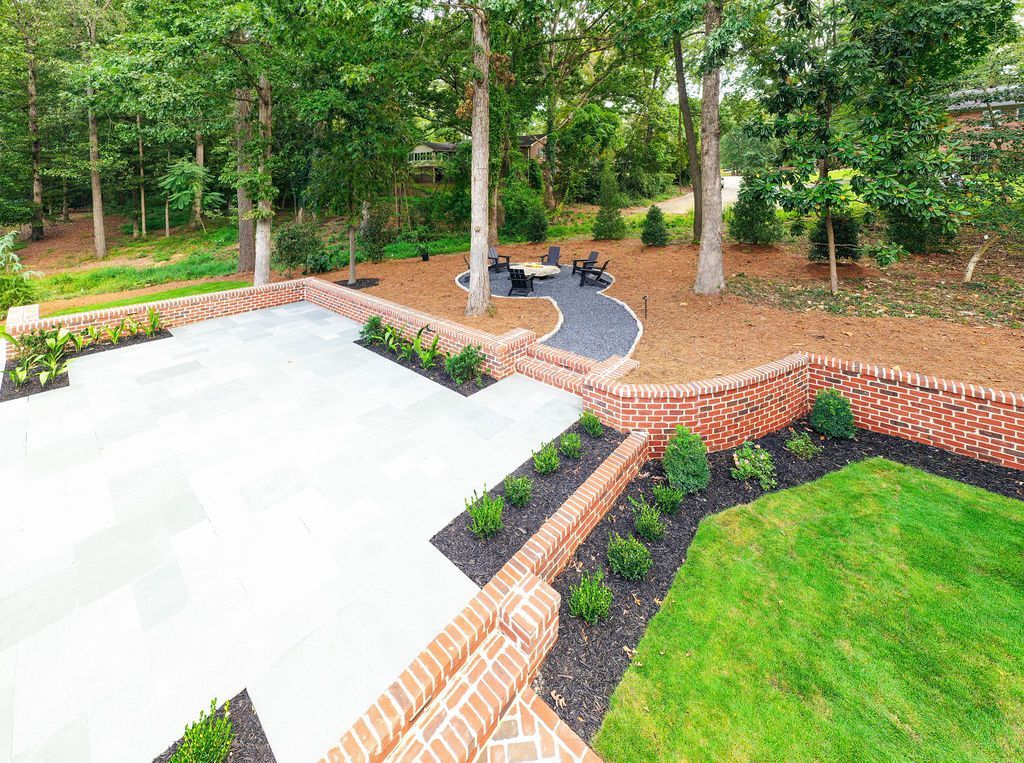 Brick garden patio with stone seating, potted plants, and green lawn beside a wooded area
