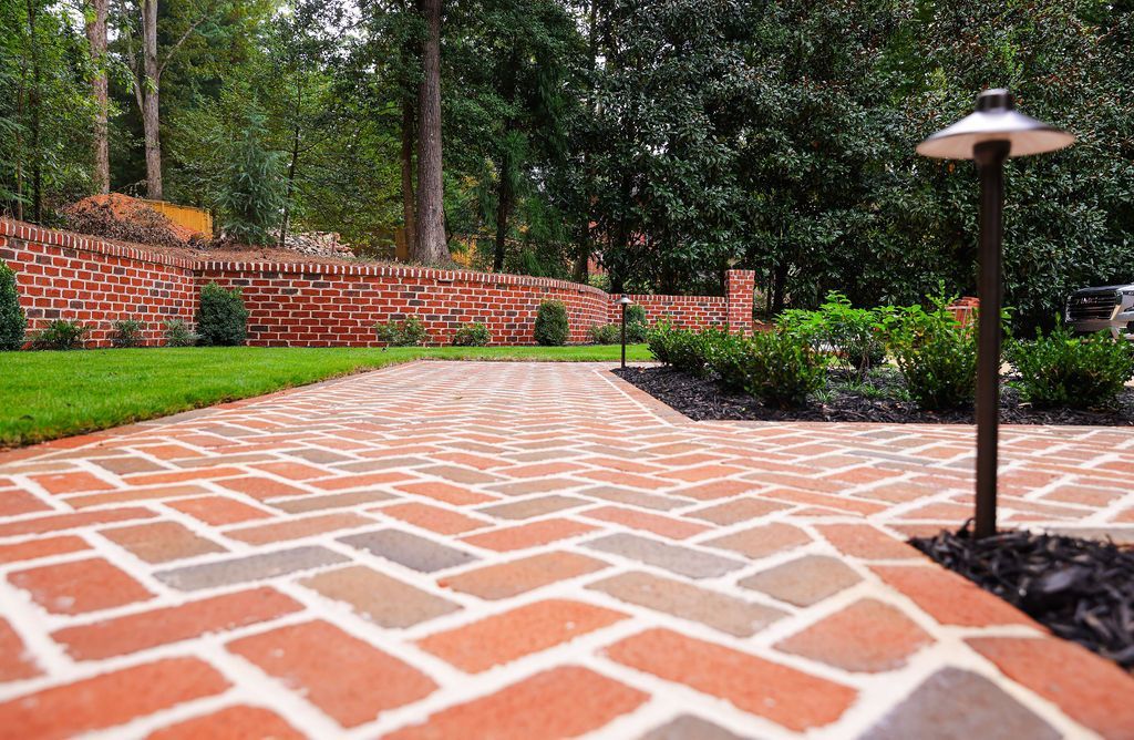 Brick patio walkway with white mortar, bordered by greenery, a brick wall, and a black lamp post.