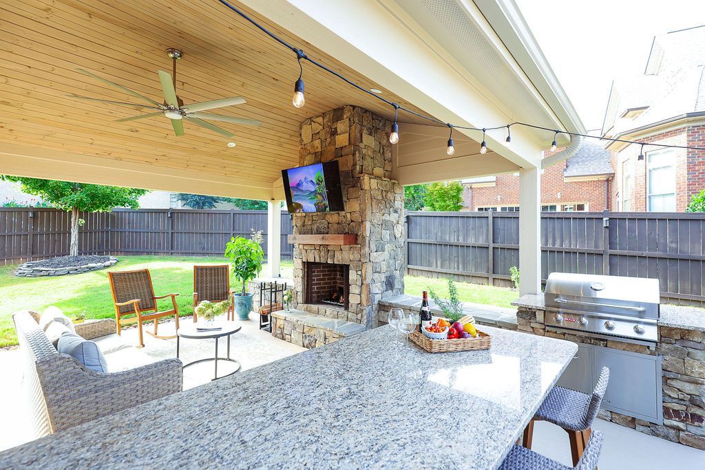 Covered outdoor patio with granite counter, grill, TV, seating, and a fire pit overlooking a fenced yard