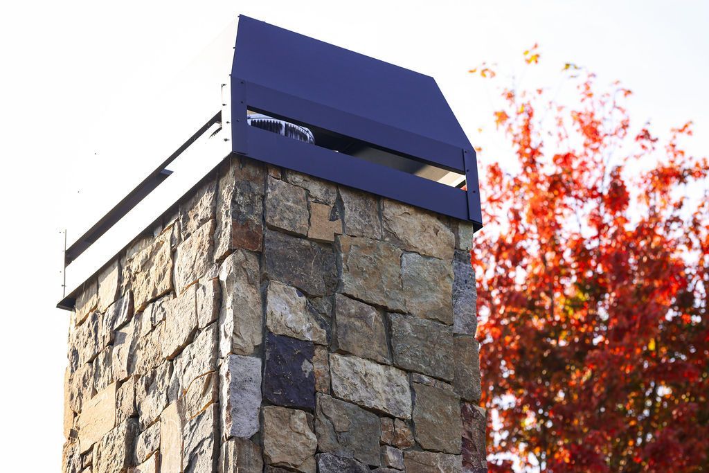 Stone chimney with a black metal cap against autumn trees and bright sky