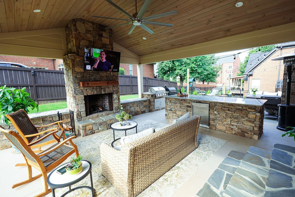 Covered patio with wicker seating, stone fireplace, and TV in a backyard.