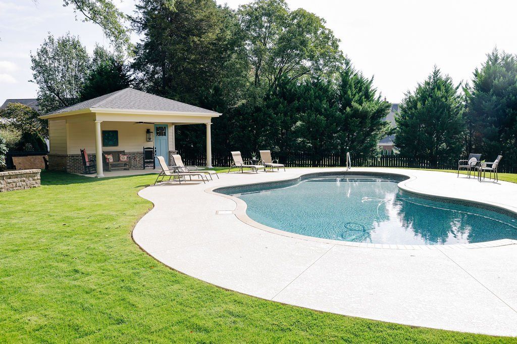 Backyard pool with curved deck, lawn, lounge chairs, and a covered patio surrounded by trees