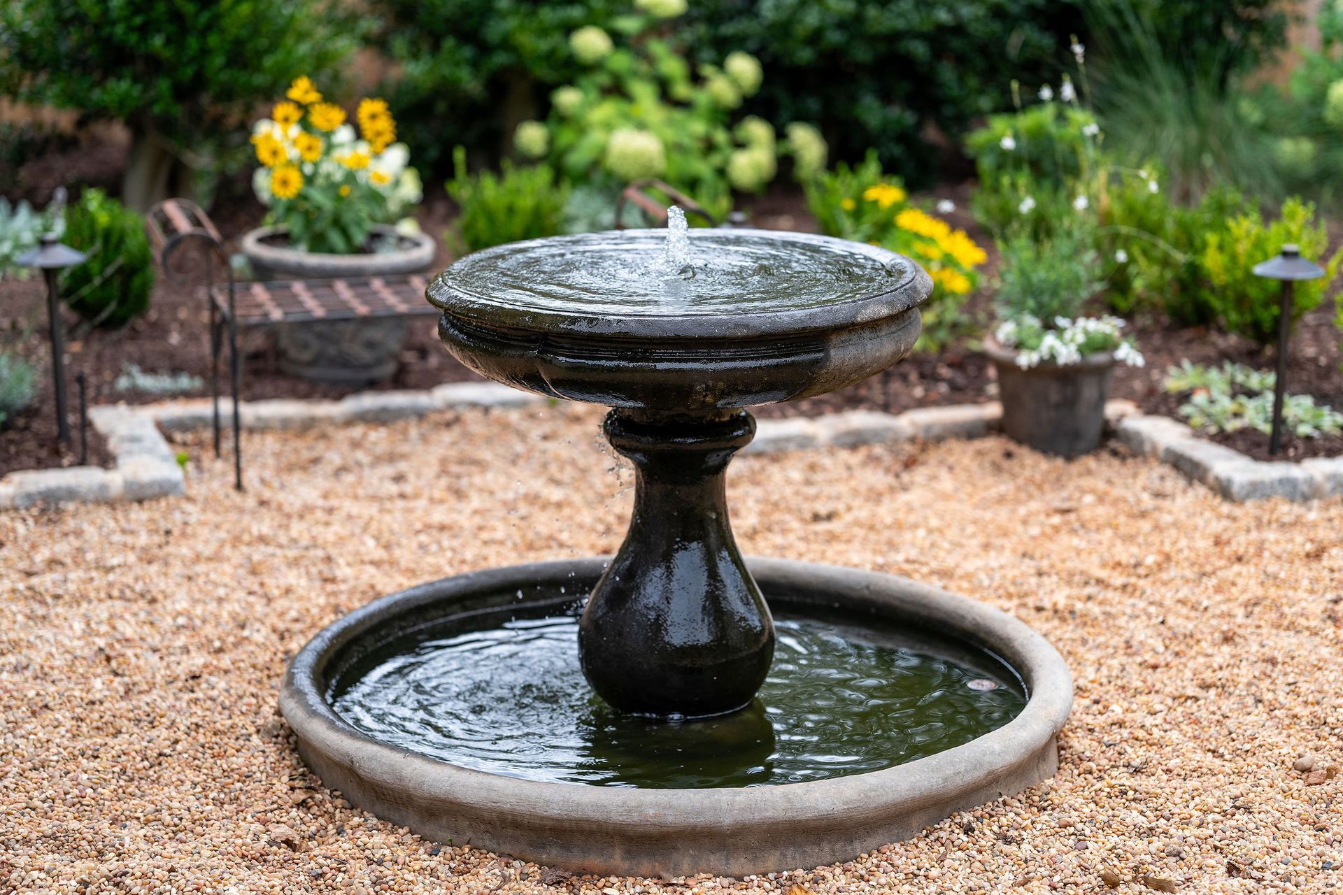 Tiered stone birdbath in a garden, with water ripples and yellow flowers in the background