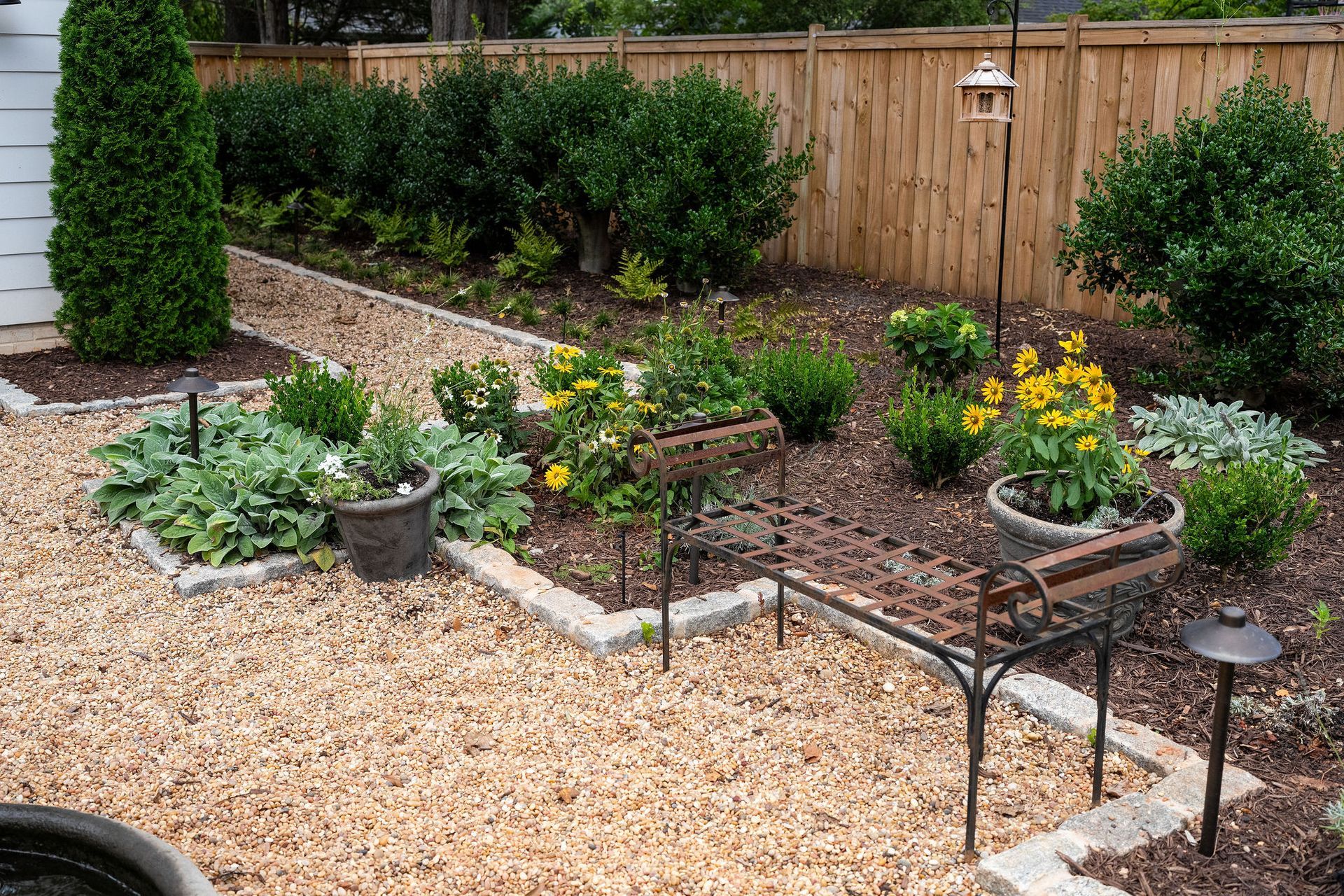 Landscaped backyard garden with gravel paths, granite cobble border, yellow flowers, and a wooden fence