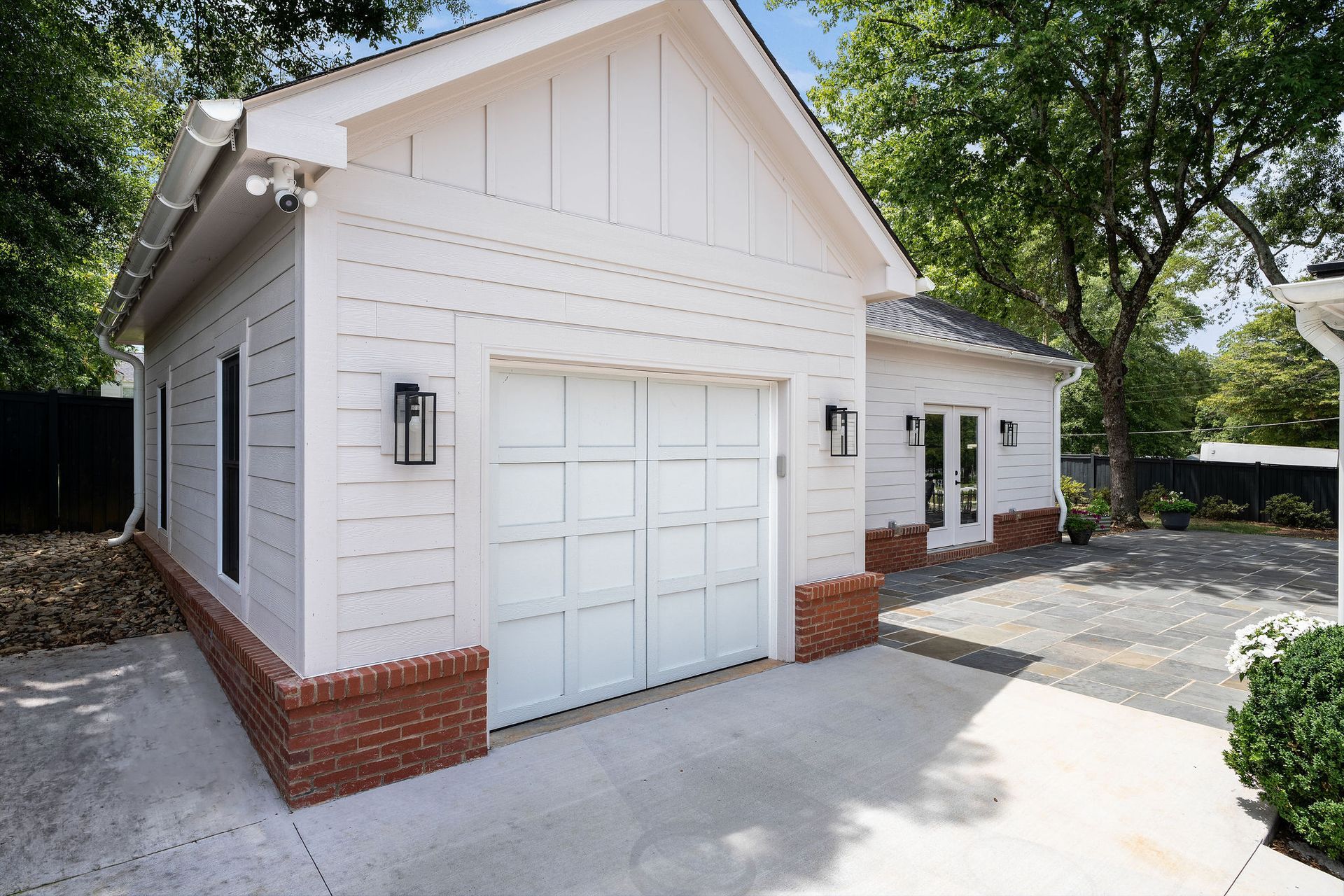 White detached garage with double doors and brick base, surrounded by trees and a paved driveway.