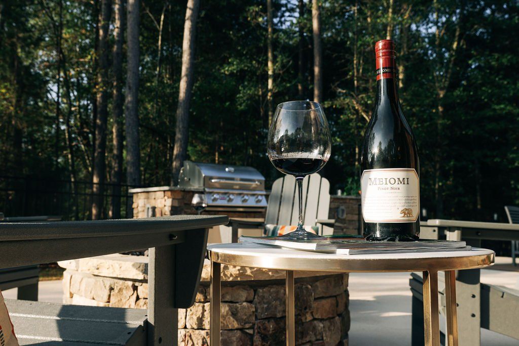 Wine bottle and glass on an outdoor table with a barbecue grill in a wooded backyard