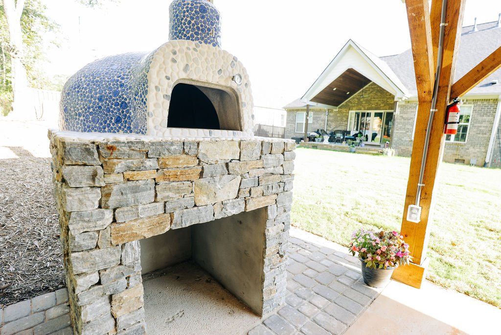 Stone outdoor pizza oven on a wooden deck beside a covered patio, with a small flower pot nearby.