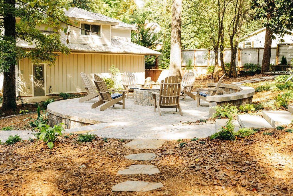 Backyard patio with stone seating area, chairs, and a stepping-stone path beside a house and trees