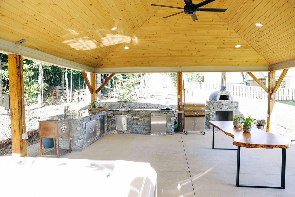 Open-air patio with wooden roof, stone bar, grill, and tables by a bright poolside area