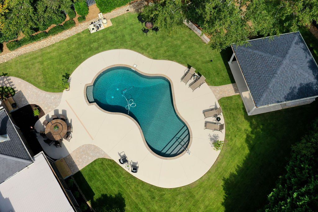 Aerial view of a backyard pool with tan deck, lounge chairs, lawn, and surrounding houses.
