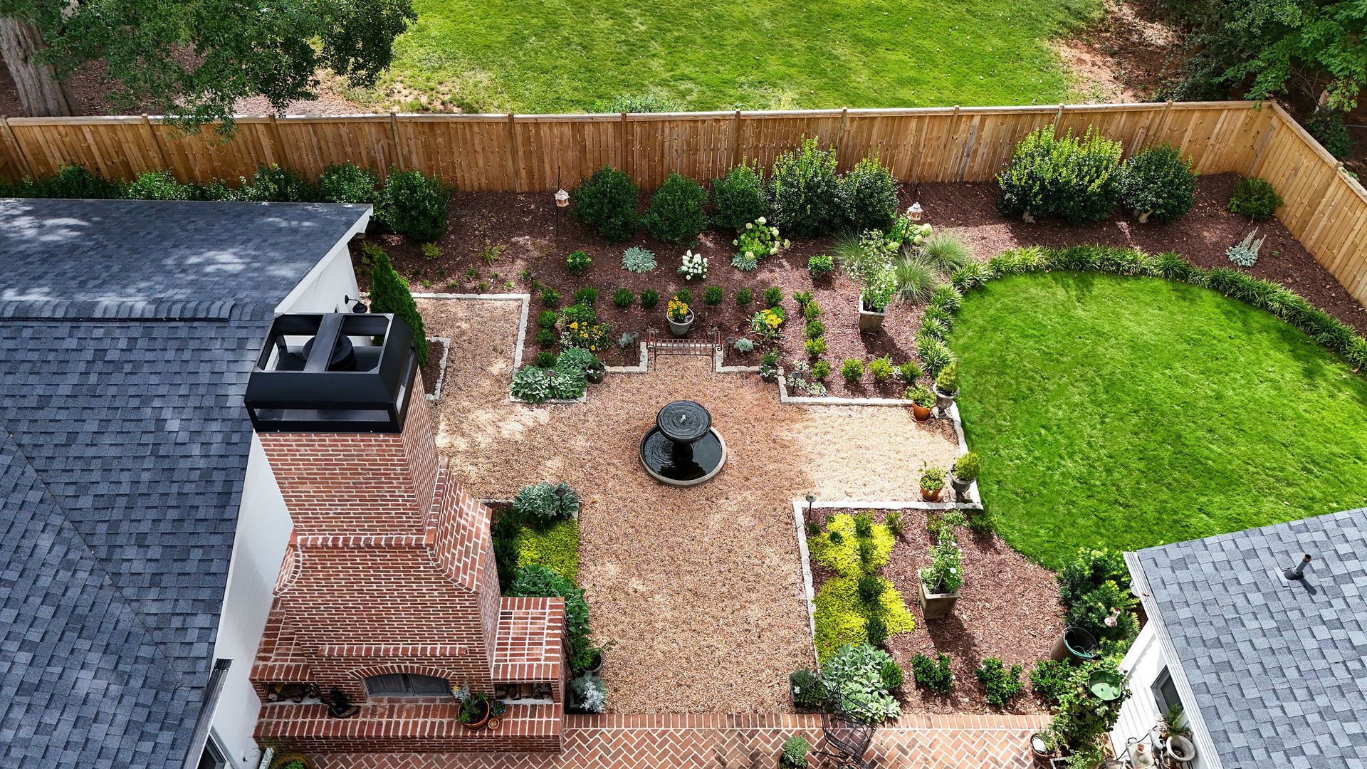 Backyard garden with a brick patio, cobble granite border, potted plants, green lawn, and brick fireplace. 