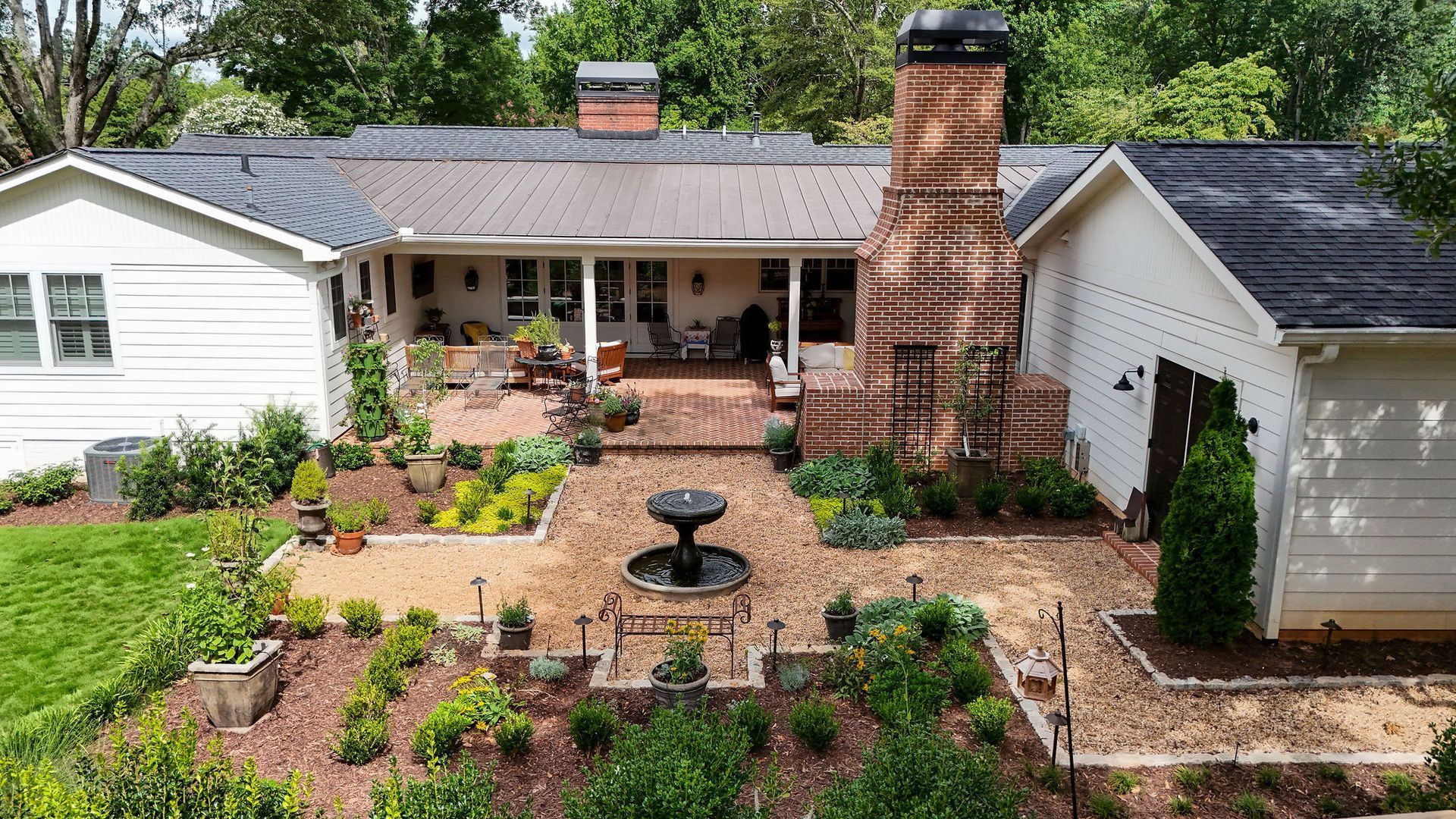 Backyard view of a white house with a brick chimney, covered back porch, and landscaped garden path with a fountain.