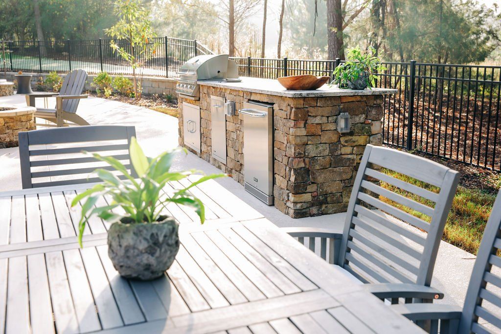 Outdoor patio with a white table, chairs, potted plant, and stone grill area in sunlight