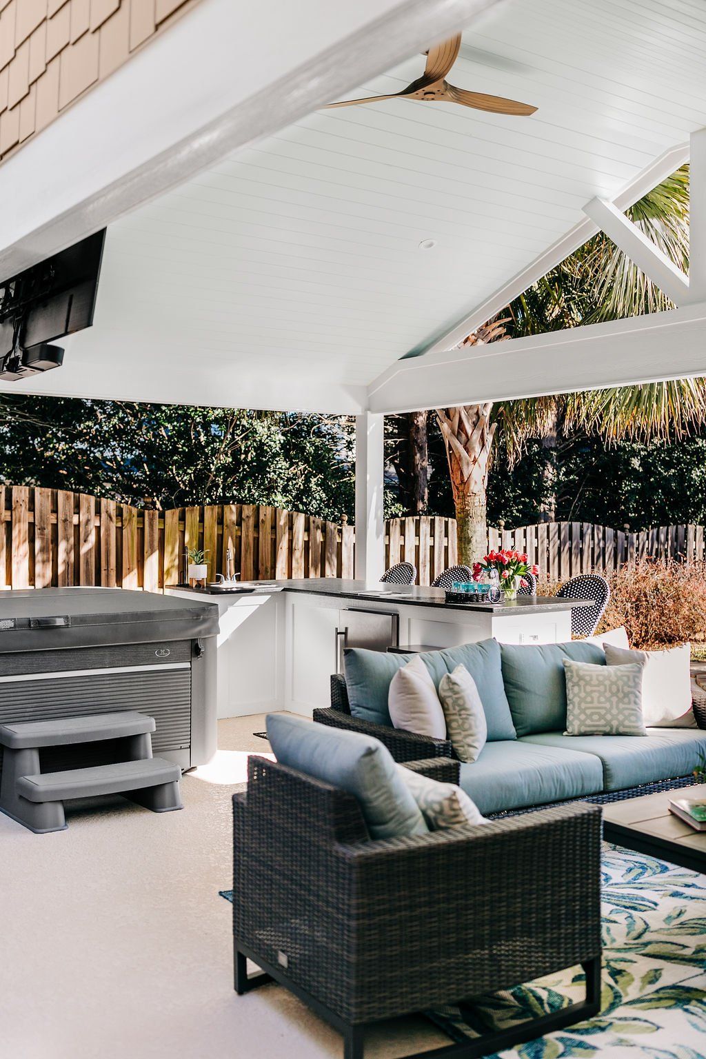 Covered patio with gray wicker seating, built-in grill, and white roof beams overlooking a wooden fence.