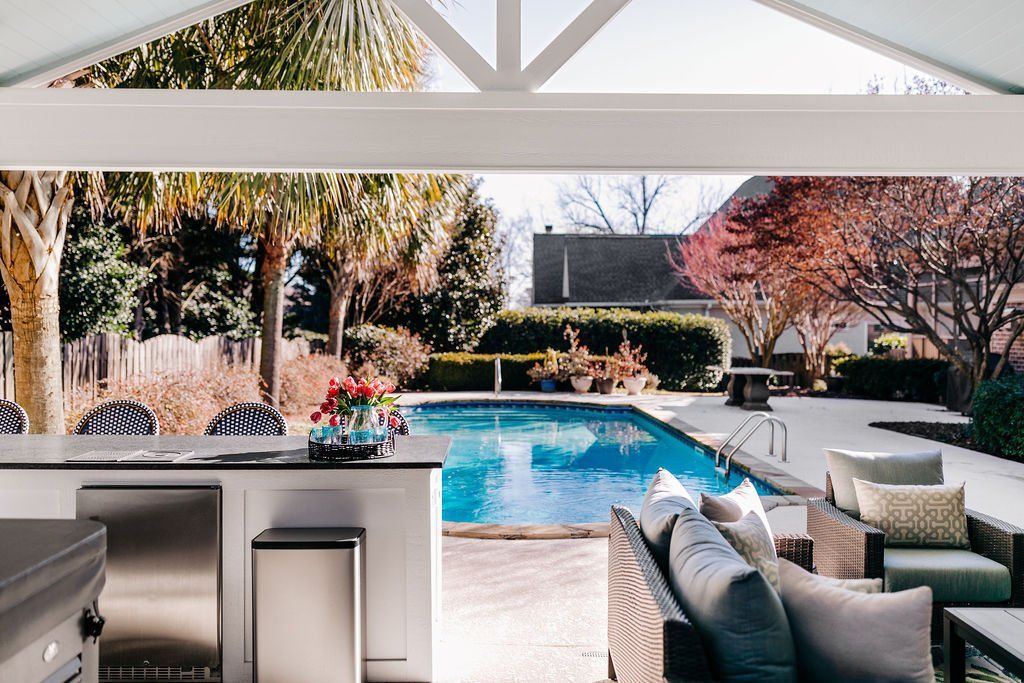 Patio overlooking a blue pool, with outdoor seating and bar stools, framed by palm trees and a sunny backyard.