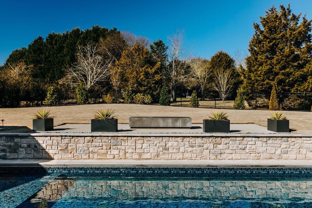 Outdoor pool with stone deck, planters, and trees under a clear blue sky