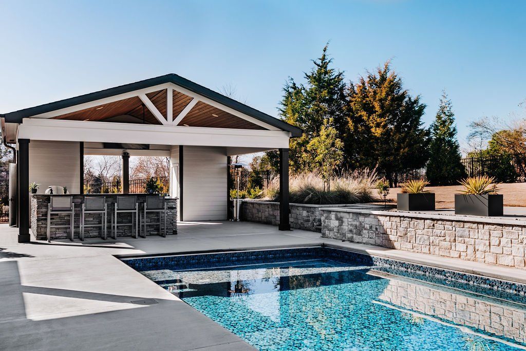 Backyard pool beside a covered patio with lounge chairs and stone retaining wall
