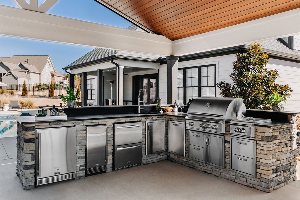 Outdoor stainless-steel grill kitchen under a patio, with a built-in barbecue, sink, and storage cabinets