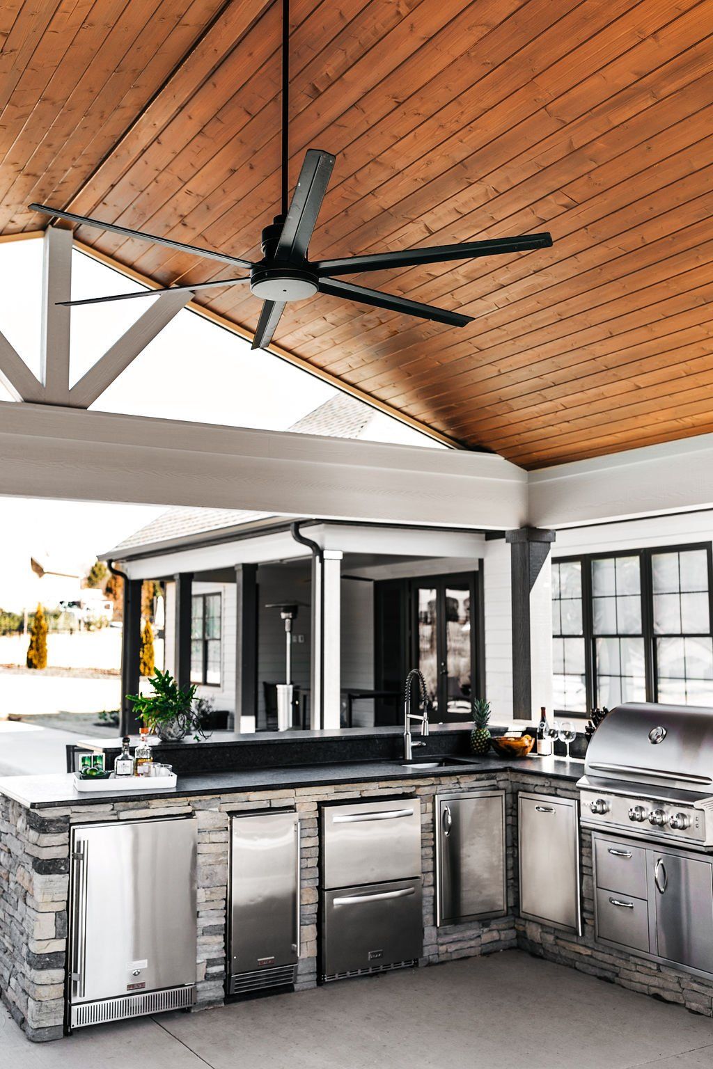 Outdoor kitchen with stainless steel grill under a wood-paneled ceiling and black ceiling fan