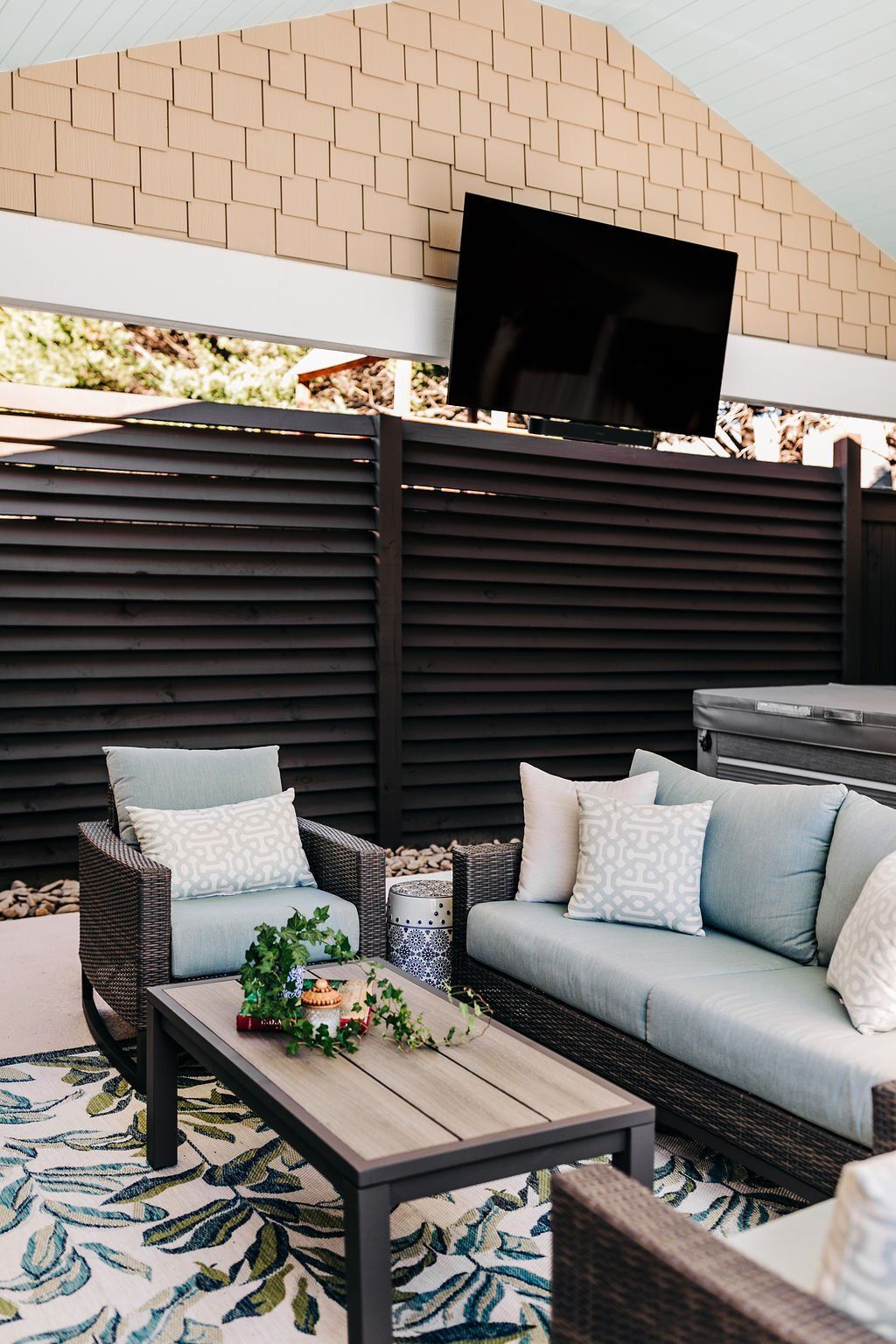 Outdoor patio seating with gray sofas, coffee table, and wall-mounted TV behind a slatted privacy screen