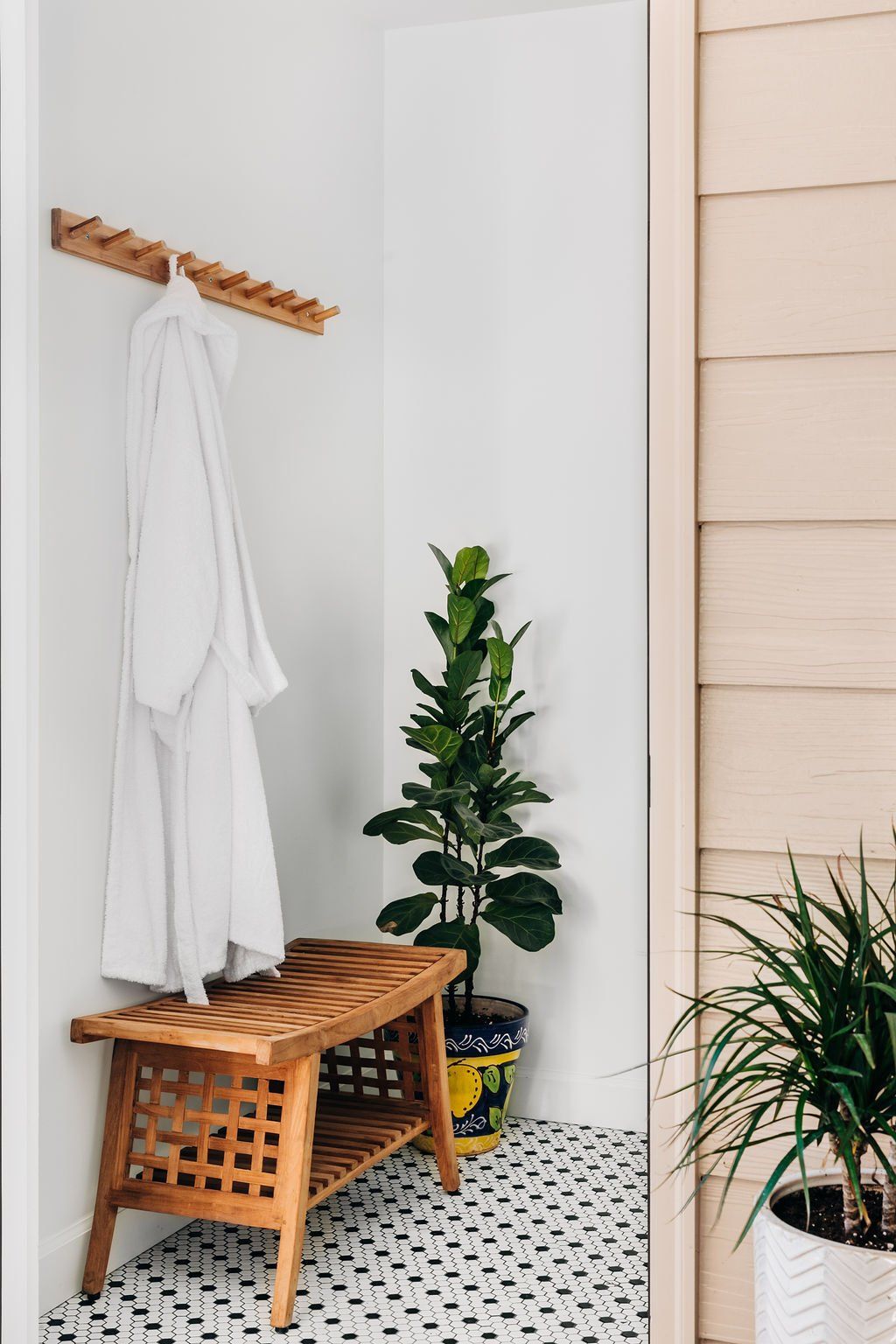 White robe on wooden coat rack beside bench and potted plants in a bright tiled entryway