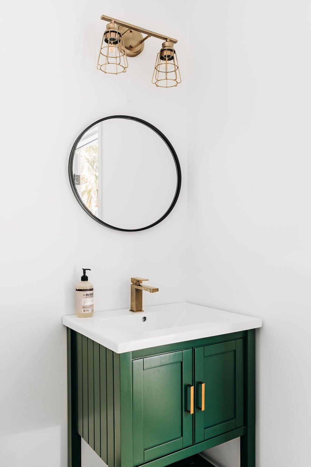 Small bathroom vanity with green cabinet, round mirror, brass faucet, and wall sconce lighting.