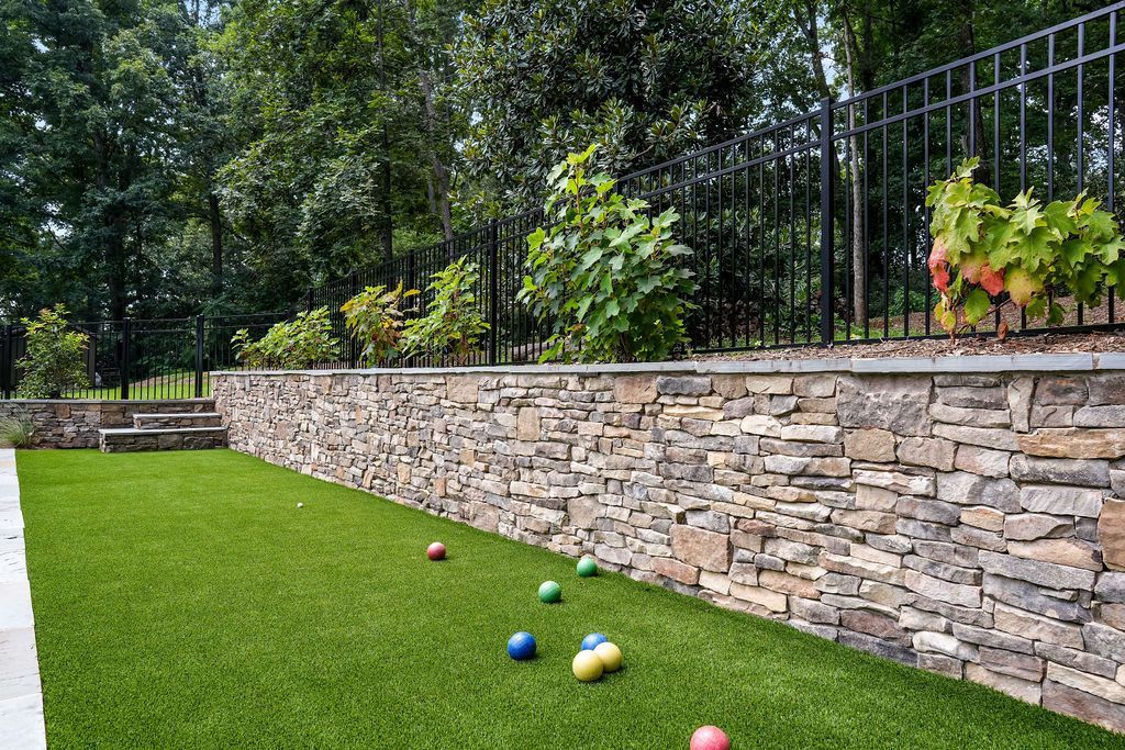 Backyard artificial turf with colorful balls by a stone wall and black fence under trees